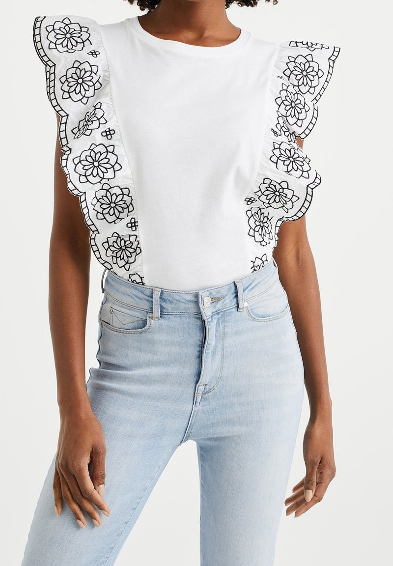 Vit t-shirt med stora svarta blomsterbroderade volanger längs ärmarna, kombinerad med ljusblå högmidjade jeans.