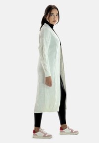Larga chaqueta de punto blanca con detalles de trenzado, llevada sobre un cuello de tortuga negro, combinada con leggings negros y zapatillas con detalles rosas.