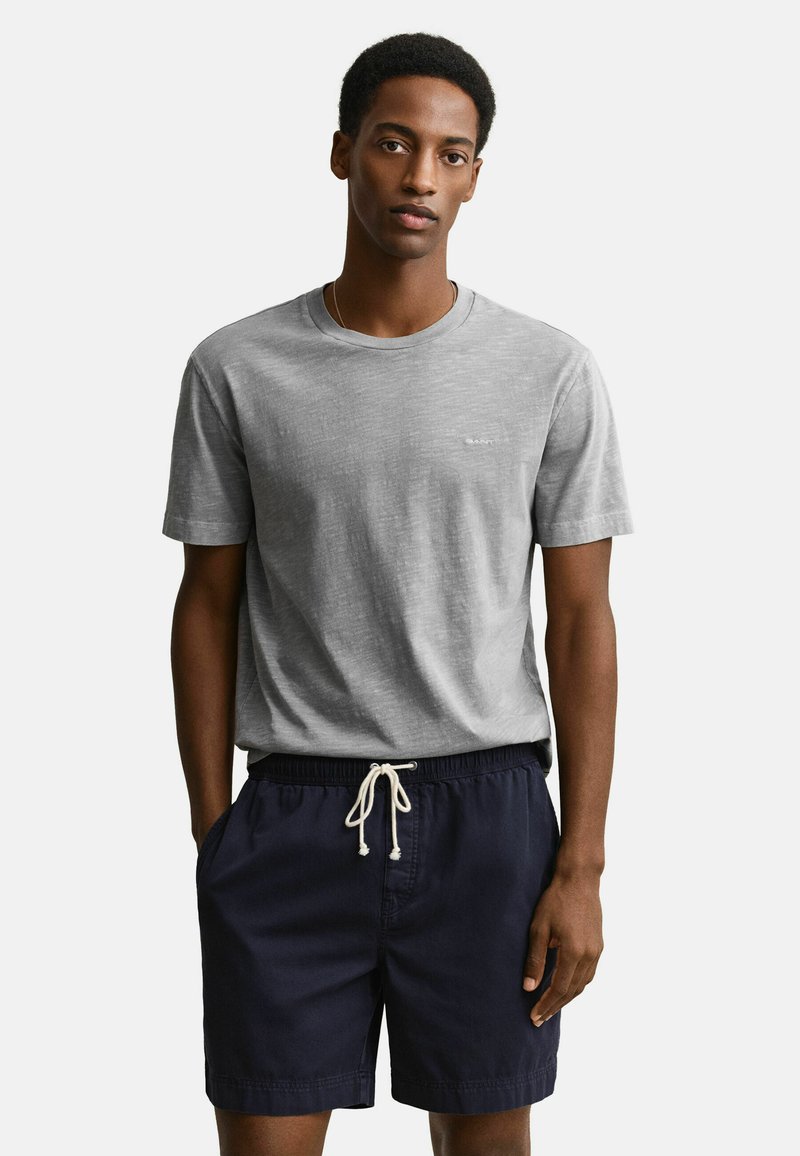 Jeune homme portant un t-shirt à manches courtes gris clair et un short à cordon de serrage bleu marine foncé, debout avec une main dans la poche.