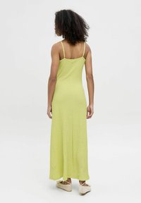 Robe longue et texturée de couleur vert lime avec des bretelles fines, une coupe décontractée et une silhouette fluide, associée à des chaussures légères à enfiler en osier.