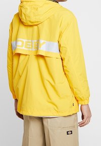 Yellow lightweight jacket featuring a hood, white striped detailing, and a bold logo on the back. Smooth texture and relaxed fit.