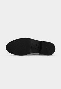 Semelle de chaussure noire avec une prise texturée à l'avant et au talon, des coutures visibles et une marque rectangulaire près de la zone de la voûte plantaire.
