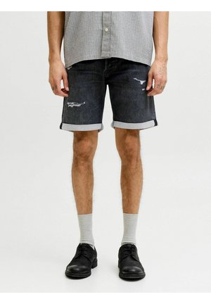 Muž nosí čierne rifľové šortky vyhrnuté hore s miernym opotrebovaním, sivé ponožky, čierne spoločenské topánky a sivú károvanú košeľu.