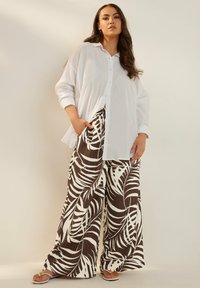 Valkoinen nappikauluspaita yhdistettynä leveälahkeisiin housuihin, joissa on ruskeita ja valkoisia trooppisia lehtikuvioita. Silkkinen rakenne, rento istuvuus.