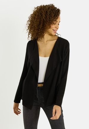 Mujer con cabello rizado que lleva un cárdigan negro abierto al frente, top blanco, jeans oscuros y un cinturón con ojales dorados, mirando hacia un lado y sonriendo.