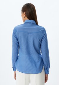 Camisa a rayas azules con cuello estructurado, mangas largas y puños abotonados. Presenta un diseño de yugo en la parte trasera. Textura de tela suave.