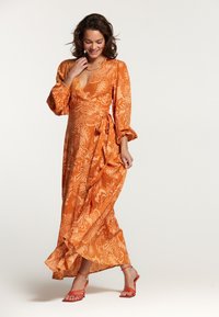 Lang, wikkeldraag jurk in fel oranje met een bloemenpatroon, voorzien van lange puffmouwen en een getailleerde taille. Gepaard met sandaaltjes met bandjes.