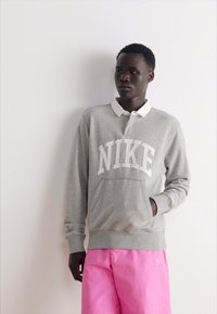 Siv sweatshirt z belim logotipom "NIKE", s kapuco in kengrovo žepom, v kombinaciji z roza kratkimi hlačami. Gladek bombažen material, sproščen kroj.