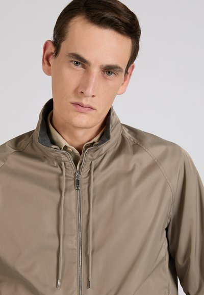 Giovane uomo con capelli castani corti che indossa una giacca marrone chiaro con cerniera sopra una camicia beige con collo, rivolto in avanti con espressione neutra.