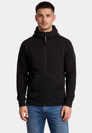 Mężczyzna ubrany w czarną bluzę z kapturem zapinaną na zamek oraz niebieskie dżinsy, stojący na jednolitym jasnym tle.