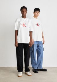 Bílé grafické trička s červeným logem Calvin Klein, spojené s černými a modrými širokými kalhotami. Viditelné neformální obuvi.