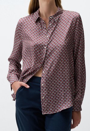 Femme portant une chemise à manches longues boutonnée avec un motif géométrique en losanges rouge, blanc et bleu et un pantalon bleu foncé, main dans la poche.