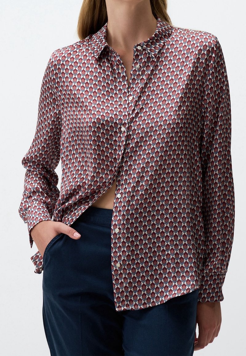 Femme portant une chemise à manches longues boutonnée avec un motif géométrique en losanges rouge, blanc et bleu et un pantalon bleu foncé, main dans la poche.