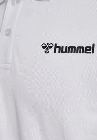 Biała koszulka polo z czarnym logo hummel i ikonką pszczoły na piersi, z trzema białymi guzikami przy kołnierzyku.