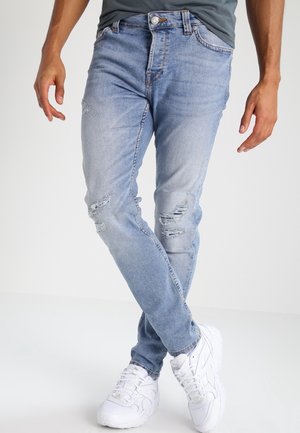 Man wearing light blue ripped jeans and white sneakers, standing with legs crossed and arms relaxed by his sides.