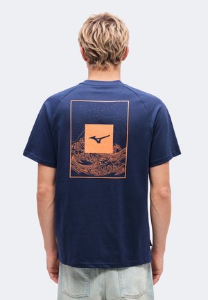Une personne vue de dos porte un T-shirt bleu marine orné d’un motif de vague orange et d’un logo dans le dos, associé à un short en jean clair.