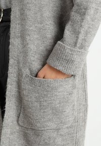 Grå cardigan av mjuk, texturerad ull med lång design, ribbade ärmslut och sidofickor. Enkelt och funktionellt stil.