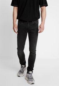 Jeans negros de corte ajustado con una textura desteñida, cinco bolsillos y detalles de costura sutiles, combinados con zapatillas deportivas grises.