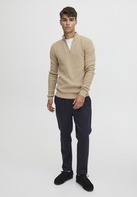 Beige strikkegenser med høy krage og kvart glidelås, paired med mørkeblå bukser og svarte sko, som skaper et avslappet men likevel strukturert utseende.