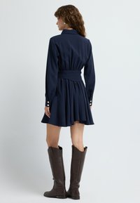 Granatowa sukienka z długimi rękawami, dopasowaną talią, rozkloszowanym dołem i złotymi guzikami. W połączeniu z wysokimi brązowymi skórzanymi butami.