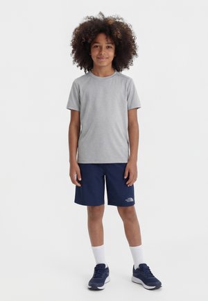 Otrok s kodrastimi lasmi stoji, oblečen v sivo majico, mornarsko modre kratke hlače, bele nogavice in mornarsko modre superge na enostavni belem ozadju.