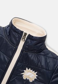 Navy blue puffer jacket with quilted texture, cream interior lining, and a front zipper featuring a logo appliqué.