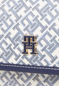 Hvid taske med blåt geometrisk mønster, marineblå detaljer og guld logo dekoration. Struktureret materiale og syninger fremhæver designet.