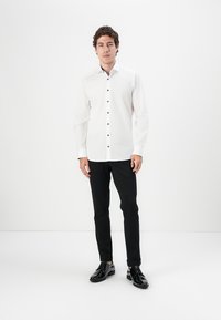 Camisa blanca de vestir con botones negros, de manga larga y cuello estándar, combinada con pantalones negros y zapatos negros brillantes.