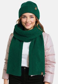 Zielona dzianinowa czapka beanie i pasujący szalik, prążkowana textura, z drobnym detalem metki. Noszone z jasnoróżową kurtką puchową i białym topem.