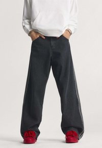 Voľné čierne denimové nohavice so sivými pásikmi na bokoch, s vysokým pásom a širokými nohavicami. Oblečené s jasne červenými teniskami a bielou mikinou.