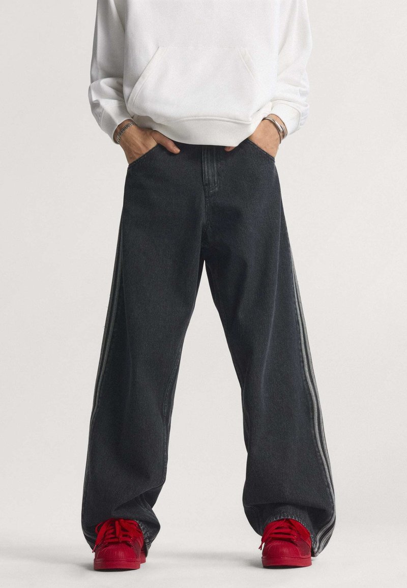 Locker sitzende schwarze Denim-Hosen mit Seitenstreifen, ausgestattet mit hohem Bund und weitem Bein. Kombiniert mit leuchtend roten Sneakern und einem weißen Hoodie.