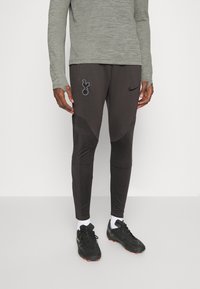 Pantalons de sport Nike en gris foncé avec un design texturé, des accents de logo et une coupe ajustée ; associés à des chaussures de sport noires et des chaussettes blanches.