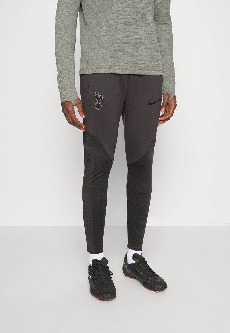 Pantalons de sport Nike en gris foncé avec un design texturé, des accents de logo et une coupe ajustée ; associés à des chaussures de sport noires et des chaussettes blanches.