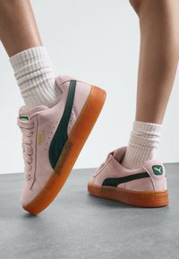 Baskets en suede rose avec des accents verts, dotées d'une semelle en caoutchouc orange texturée et d'un col rembourré. Des chaussettes blanches mi-mollet sont portées.