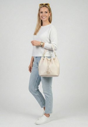 Smilende kvinde med langt blondt hår iført hvid sweater, lyseblå jeans, hvide sneakers, gule solbriller og bærer en beige håndtaske over skulderen.