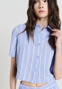 Camicia button-up azzurro chiaro con righe bianche verticali, maniche corte e lunghezza accorciata. Presenta un colletto a punta e una texture sottile.