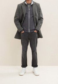 Cappotto di lana grigio su una felpa con zip grigio scuro, jeans neri e sneakers bianche. Dettaglio a righe sulla felpa, vestibilità classica.