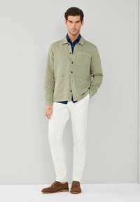 Giacca verde chiaro con chiusura a bottone e due tasche frontali, abbinata a una camicia blu navy e pantaloni bianchi. Scarponi in suede marrone completano il look.