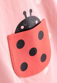 Τσέπη από κόκκινο ύφασμα με σχέδιο πασχαλίτσα και μαύρα στίγματα πάνω σε ροζ ύφασμα, που δείχνει το κεφάλι μιας καρτούν πασχαλίτσας με κλειστά μάτια να κοιτάζει έξω από την τσέπη.