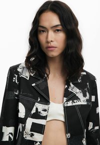 Black jacket with white graphic print, featuring a notched lapel and contrasting stitching. Material appears textured and lightweight.
