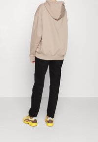 Persona con felpa beige, pantaloni neri e sneakers gialli in piedi, di fronte a un muro bianco su un pavimento chiaro.