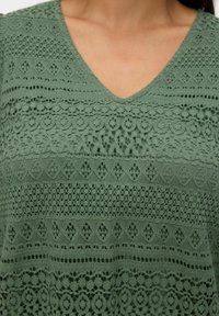 Femme portant un haut vert en V avec des motifs de dentelle horizontaux détaillés et des designs de découpes géométriques.