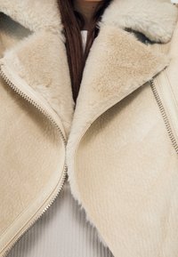 Béžová bunda z umělé kožešiny se širokým límcem, viditelným stříbrným zipem a měkkou texturou. Pod ní je částečně viditelný světle šedý žebrovaný top.