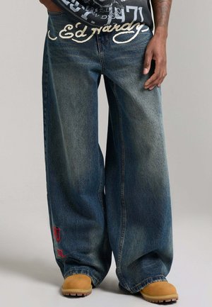 Szerokie, wyblakłe niebieskie dżinsy z czerwonym haftem przy krawędzi, noszone z beżowymi butami roboczymi i czarną koszulką z nadrukiem włożoną do środka.