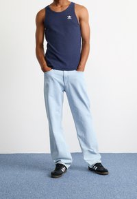 Navy ribbed tank top with white logo, light blue loose-fit jeans, and black sneakers with white stripes. Model poses with hands in pockets.