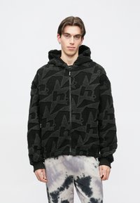 Jauns vīrietis melnā teksturētā kapucjakā ar abstraktiem rakstiem un gaišām krāsotām biksēm, stāv pret vienkāršu baltu fonu.