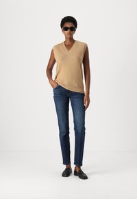 Beige ärmelloser Strickpullover mit V-Ausschnitt, kombiniert mit dunkelblauen eng anliegenden Jeans und schwarzen Slippern. Das Modell steht mit einer Hand in der Tasche.