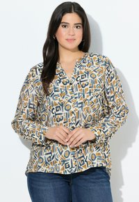 Eine Frau mit langen dunklen Haaren, die goldene Creolen trägt und eine beigen Knopfleiste-Bluse mit abstraktem blauem und braunem Muster anhat, kombiniert mit blauen Jeans.