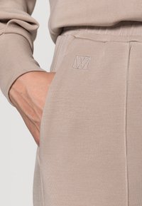 Ljust beige byxor gjorda av mjukt tyg, med en sidficka och diskret broderad logotyp. Resårmidja med sytt detaljer.