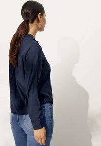 Marineblauwe blouse met lange mouwen en een ruches decolleté, gedragen met strakke blauwe jeans, die een soepele textuur en een relaxte pasvorm tonen.